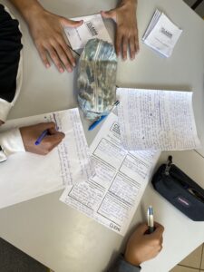« Trois personnes travaillent autour d’une table sur des documents scolaires : feuilles manuscrites remplies de notes en bleu, fiches imprimées avec des questions, et petits livrets intitulés “CLASSE INVESTIGATION” et “CLEMI”. Deux trousses, l’une camouflage et l’autre noire, sont posées sur la table. Les mains des participants écrivent ou relisent leurs réponses, suggérant une activité de groupe ou un exercice pédagogique. 