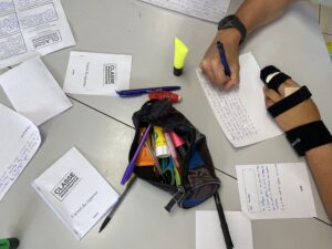 Une personne portant une orthèse au poignet écrit avec un stylo bleu sur une feuille posée sur une table encombrée de documents scolaires. On distingue des papiers imprimés intitulés “CLASSE INVESTIGATION”, “Carnet du reporter” et “CLEMI”, ainsi que des notes manuscrites en français évoquant les réfugiés. Autour, une trousse ouverte contenant stylos et surligneurs, un marqueur jaune et un stylo noir. L’ensemble suggère une activité pédagogique ou journalistique en classe.